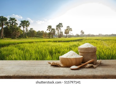 Jasmine Rice In Bowl And Burlap Sack On Wooden Table With The Rice Field Background