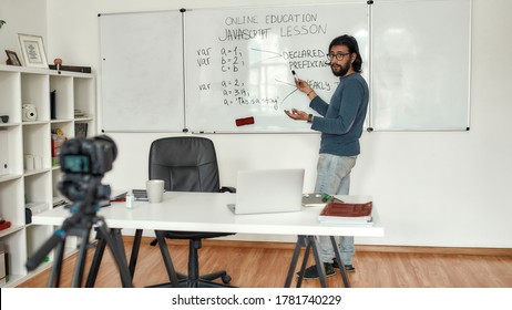 Javascript lesson. Young bearded male teacher wearing glasses pointing at whiteboard and teaching Javascript, giving lesson online. Recording video blog. Focus on a man. E-learning. Distance education