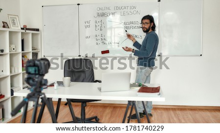 Javascript lesson. Young bearded male teacher wearing glasses pointing at whiteboard and teaching Javascript, giving lesson online. Recording video blog. Focus on a man. E-learning. Distance education
