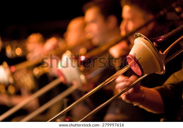Jazz Big Band trombone section with\
cup mutes. Shallow focus on the foreground trombonist with the rest\
of the section falling into background\
blur.\
