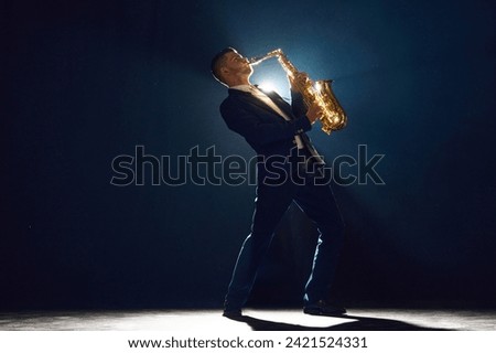 Jazz musician in suit virtuously playing saxophone stands with dramatic backlight behind him on dark stage. Concept of art, instrumental music, dance, culture, festivals and concerts.