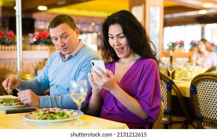 Jealous man peeking into wife smartphone, reading her messages while they have dinner in restaurant. Woman ignoring husband. Family phubbing concept..