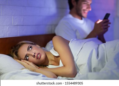 Jealous Wife Suspecting Infidelity While Husband Texting On Mobile Phone Lying In Bed At Home At Night. Low Light