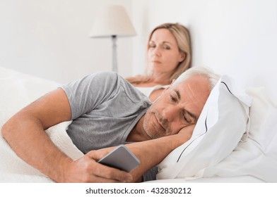 Jealous woman spying her husband mobile phone while he is reading a message. Senior couple in bed while wife is angry as husband using smartphone. Husband ignoring wife and texting on smartphone.
