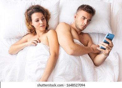 Jealous woman tracking her man in bed, spying on a cell phone while the male is reading a message. A handsome is in bed with a mobile smartphone, a female is peeping at his online communication sms.