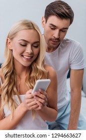 jealous young man looking at beautiful smiling wife using smartphone in bedroom 