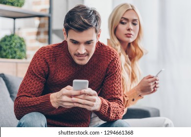 jealous young woman with smartphone looking at smiling boyfriend using smartphone at home, relationship problem concept