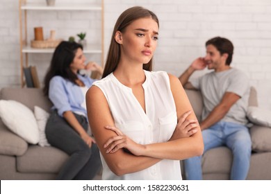 Jealousy Concept. Offended Girlfriend Standing Crossing Hands While Her Boyfriend Flirting With Girl Sitting On Sofa Indoor. Shallow Depth - Powered by Shutterstock