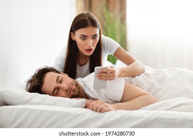 Jealousy Problem. Shocked Girlfriend Reading Messages On Cheating Husband's Phone Suspecting Infidelity In Bedroom At Home. Jealous Girlfriend Checking Boyfriend's Smartphone. Selective Focus