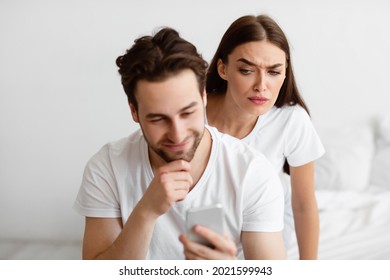 Jealousy. Suspicious Wife Reading Husband's Messages On Smartphone Standing Behind His Back While He Texting In Social Media Indoors. Jealous Girlfriend Checking Boyfriend's Chats Suspecting Affair