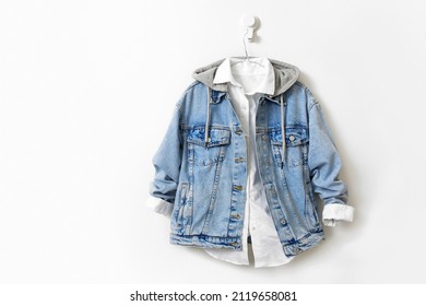 Jean Jacket Over White Shirt Hanging On The White Wall Empty Space. Beautiful Trendy Clothes. Unisex Clothing. Blue Denim Jacket, Coat.Vintage Style Apparel.