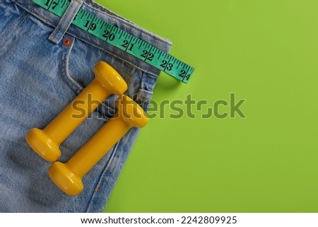 Jeans, dumbbells and measuring tape on light green background, flat lay with space for text. Weight loss concept