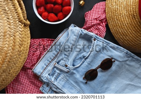 Jeans, gingham top, straw bag and hat, accessories and bowl of strawberries on dark background. Spring or summer picnic outfit. Top view.