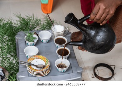 A jebena sits on a grass-covered table with small, handle-less cups arranged around it. Fresh roasted coffee beans, a mortar and pestle, 
incense smoke create a traditional Ethiopian coffee ceremony - Powered by Shutterstock