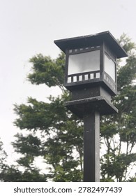 Jember, East Java, Indonesia - May 18 2026: A classic black square garden lamp post standing against lush green trees in an outdoor park.