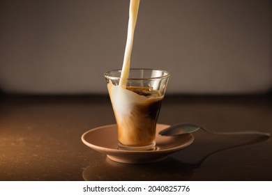 Jet of milk that falls from above fills the cup of coffee that is on top of the coffee plate, the spoon is flat on the coffee plate with a warm atmosphere and a gray background with brown soil.
