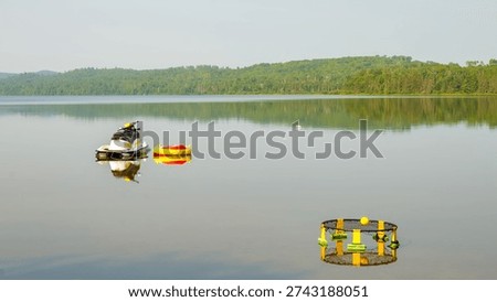 Jet ski and bright inflatable floatation device rest on calm lake water, surrounded by lush green forest and clear blue sky in the background