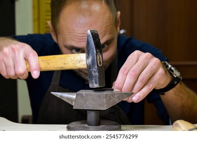 A jeweler carefully hammers metal on a small anvil in a workshop. His intense focus highlights the precision required for jewelry crafting, emphasizing the artistry in shaping metal by hand. - Powered by Shutterstock