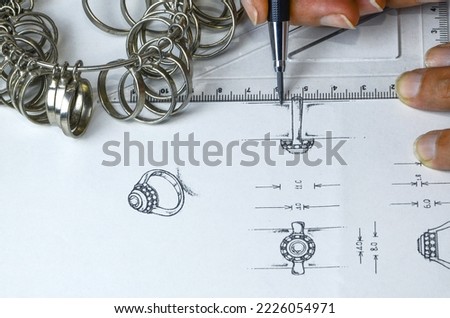 Jewelery designer drawing a diamond and gemstone ring showing the side, top, front, and imaginary full-size details with pencil and tools on a piece of paper before modeling it.
