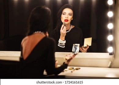Schmuck- und Beauty-Konzept - schönes Brunette Lady Portrait in modernen Schmuckstücken und schwarzes Kleid im Studio. Die Frau sitzt am Schminkspiegel und zieht einen roten Lippenstift an