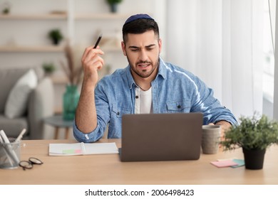 Jewish Business Guy Working On Laptop Sitting At Desk At His Workplace Indoors, Making Video Call With Clients Or Watching Webinar. Male Worker Having Online Interview Or Meeting, Holding Pen