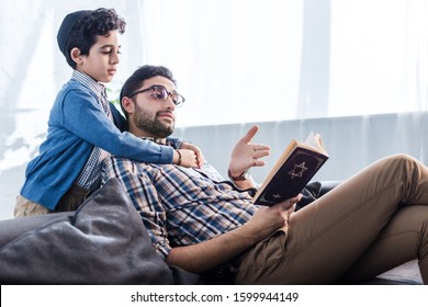 jewish father pointing with hand at tanakh and talking with son in apartment 