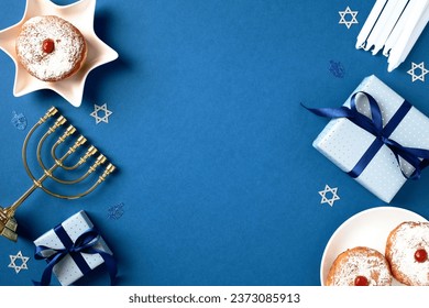 Jewish holiday Hanukkah concept. Frame of traditional jelly donuts, menorah, candles and gift boxes on blue background. Flat lay, top view.
