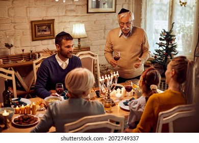 Jewish multi-generation family having a meal at dining table on Hanukkah. Focus is on senior man proposing a toast.