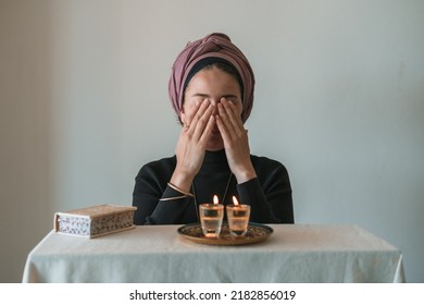 Jewish woman prays over lit Shabbat candles, covering her face with her hands. Nearby lies a religious prayer book. Jewish religious traditions