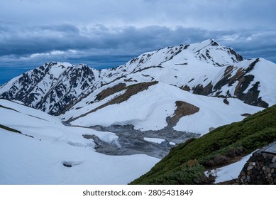 Jigokudani under a brooding storm sky — dark clouds, drifting steam and snow peaks in dramatic mountain weather.