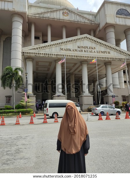 Jilbab in front of federal\
court in Kuala Lumpur Malaysia parking con with car park at the\
back high big tall building poles white in color and Malaysia\
country flags
