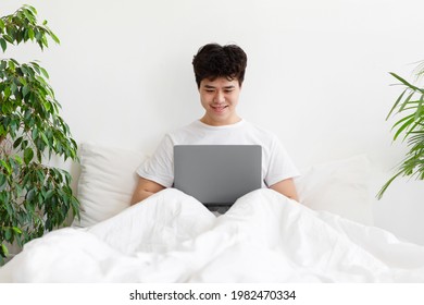 Job In Bedroom, Social Networks, Surfing, Browsing On Internet, Modem Device. Smiling Millennial Asian Man Woke Up, Sits On White Bed, Works On Laptop In Interior, On Light Wall Background, Free Space