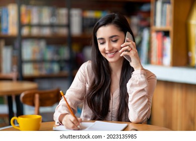 Job Hunting. Happy Arab Woman Talking By Smartphone With Hr Representetive And Taking Notes, Sitting At Cafe Interior And Smiling. Young Lady Looking For Job Opportunities