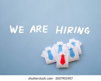 Job recruiting advertisement represented by 'WE ARE HIRING' texts on wall. One origami paper shirt and red tie is colored differently to represent the hiring position to be recruited and filled.