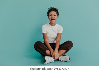 Jocund Black Girl Posing With Tongue Out On Turquoise Background. Front View Of Stunning African American Woman Sitting With Crossed Legs.