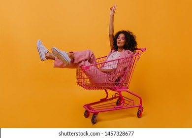 Jocund young woman with dark wavy hair enjoying photoshoot. Stylish african girl sitting in shopping cart.
