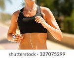 Jogger running in sportsbra with music in earphones. Midsection of determined fitness woman in sportsbra. Sporty woman is using earphones while running. She is in park on sunny day.