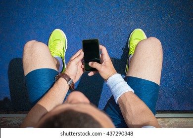 Jogger Sitting On Bench Next To The Blue Jogging Track And Using Smartphone With Blank Screen. Preparing For The Morning Workout. Top View. Technology, Sport And Fitness Concepts. 