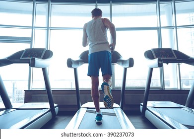 Jogging his way to good health. Full-length rear view of young man in sportswear running on treadmill in front of window at gym