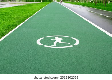 Jogging path with white runner symbol painted on green pavement, surrounded by greenery and an empty lane for cyclists, representing an outdoor fitness and recreation area