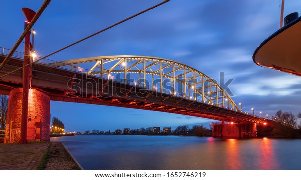 John Frost Bridge Arnhem by night. The lights are\
reflected in the water of the river, the sky turns colorful and\
then blue as clouds pass\
by.