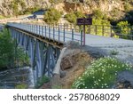 Johnsondale Bridge and 33E30 River Trail Sign over Kern River in Camp Nelson, California. Cars parked in parking lot in the background.