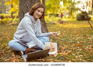 Join me. Cute girl having lunch outdoors, sitting on grass and studying on distance