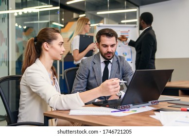 Joined Work Of Two Confident Business People In Formal Suit Working Together With Laptop At Office With Their Coworkers At Background.