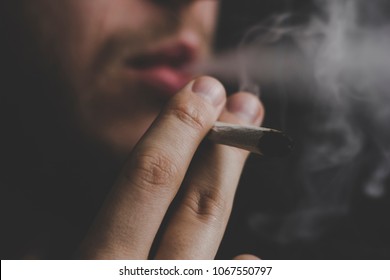 A Joint In Hand A Man Smokes Cannabis Weed, . Smoke On A Black Background. Concepts Of Medical Marijuana Use And Legalization Of The Cannabis. On A Black Background