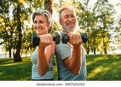 Joint workout on retirement. Beautiful and healthy elderly couple keeping dumbbells in hands while standing back to back at sunny park. Smiling family of two doing exercises for arms on fresh air. - Powered by Shutterstock