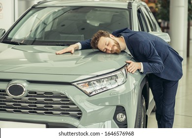 Jokey Young Caucasian Guy Hugging His New Car In Car Showroom. Happy Man Finally Gets Long-awaited Car, Wearing Formal Suit