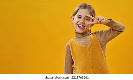 Joking caucasian little girl winking, sticking out tongue and showing victory sign. Cute female child of zoomer generation. Concept of childhood lifestyle. Yellow background. Studio shoot. Copy space - Shutterstock ID 2211768661
