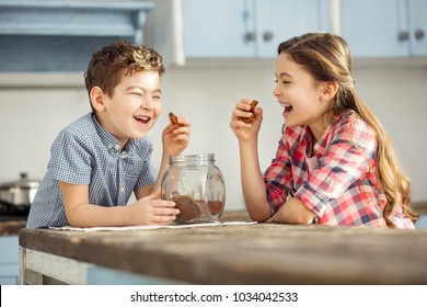 Joking. Cute Happy Little Dark-haired Brother And Sister Laughing And Eating Cookies While Sitting At The Table