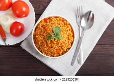 Jollof Rice, Tomatoes And Hot Peppers On A Blue Plate, Fork, Spoon On A Linen Napkin On A Brown Wooden Background. National Cuisine Of Africa.